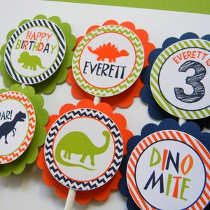 May include: Six green, orange and blue cupcake toppers with a dinosaur theme. The toppers feature dinosaurs, the words "Happy Birthday", "Roar!" and "Dino-mite", and the name "Everett" with the number "3".