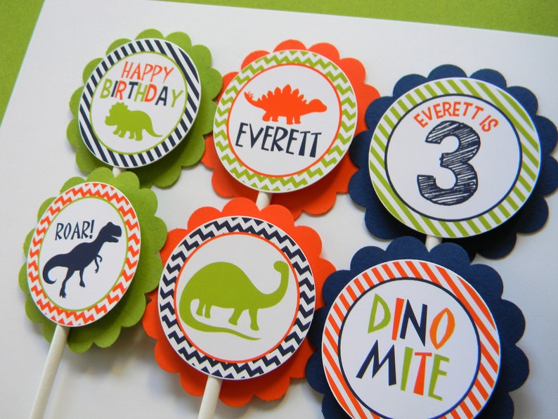 May include: Six green, orange and blue cupcake toppers with a dinosaur theme. The toppers feature dinosaurs, the words "Happy Birthday", "Roar!" and "Dino-mite", and the name "Everett" with the number "3".