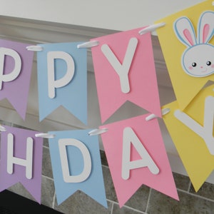 Puede incluir: Una pancarta de colores pastel con las palabras "Happy Birthday" en letras blancas. La pancarta tiene una cara de conejo linda con ojos azules y orejas rosas en una de las banderas.