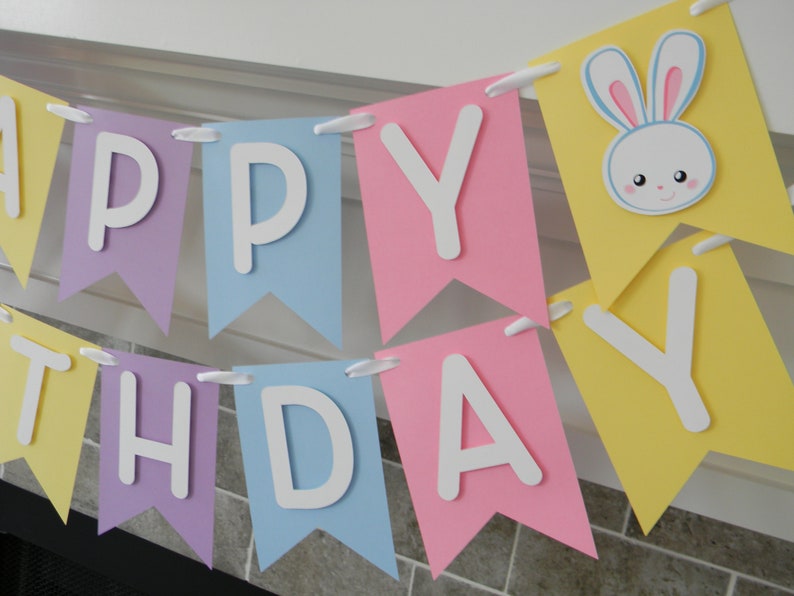 Puede incluir: Una pancarta de colores pastel con las palabras "Happy Birthday" en letras blancas. La pancarta tiene una cara de conejo linda con ojos azules y orejas rosas en una de las banderas.