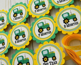May include: Green and yellow tractor themed monthly milestone baby shower decorations. The decorations feature a green tractor with black wheels and a yellow background with white polka dots. The decorations are labeled with the months of the year, from newborn to one year.