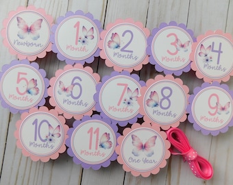 May include: A set of monthly milestone cards with a butterfly theme. Each card is round with a scalloped edge in pink and purple. The cards are labeled with months from "Newborn" to "One Year". A pink ribbon is included.