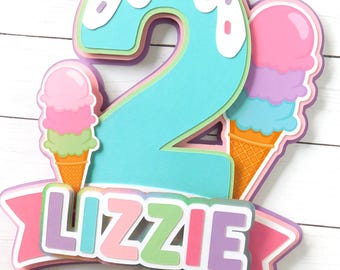 Könnte beinhalten: Bunte Tortendekoration mit der Zahl "2" in Türkis, mit einer weißen, mit Streuseln bedeckten Spitze. Zwei Eiswaffeln und der Name "LIZZIE" sind ebenfalls enthalten, ideal für eine Geburtstagsfeier.