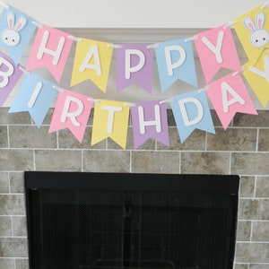 Puede incluir: Una pancarta de colores pastel con las palabras "Feliz cumplea&ntilde;os" e ilustraciones de conejos. La pancarta est&aacute; colgada sobre una chimenea negra.