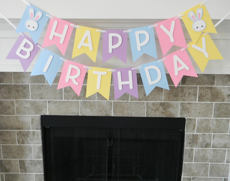 Puede incluir: Una pancarta de colores pastel con las palabras "Feliz cumplea&ntilde;os" e ilustraciones de conejos. La pancarta est&aacute; colgada sobre una chimenea negra.
