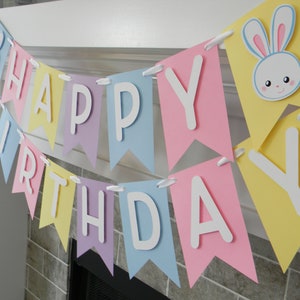 Puede incluir: Una pancarta de colores pastel con las palabras "Happy Birthday" en letras blancas. La pancarta tiene un conejito con orejas rosas en una de las banderas.