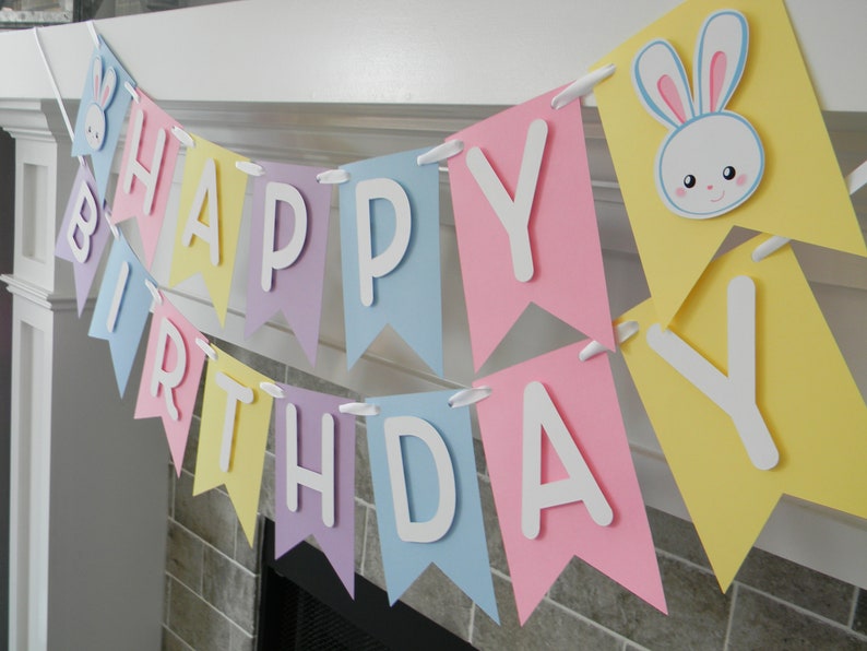 Puede incluir: Una pancarta de colores pastel con las palabras "Happy Birthday" en letras blancas. La pancarta tiene un conejito con orejas rosas en una de las banderas.