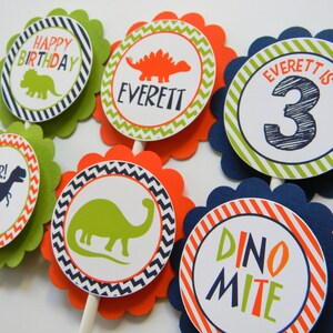 May include: Six green, orange and blue cupcake toppers with dinosaur designs. The toppers feature a dinosaur silhouette, the words "Happy Birthday", "Everett", "Roar!" and "Dino-mite", and the number "3".