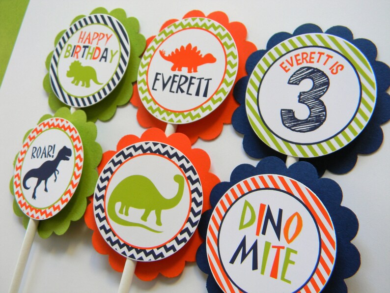 May include: Six green, orange and blue cupcake toppers with dinosaur designs. The toppers feature a dinosaur silhouette, the words "Happy Birthday", "Everett", "Roar!" and "Dino-mite", and the number "3".