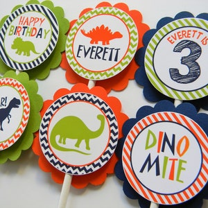 May include: Six green, orange and blue cupcake toppers with dinosaur designs. The toppers feature a dinosaur silhouette, the words "Happy Birthday", "Roar!" and "Dino Mite", and the name "Everett" with the number "3".