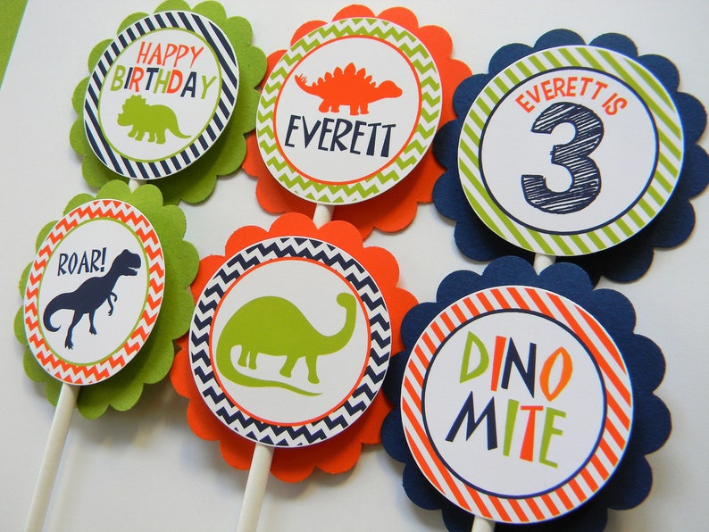 May include: Six green, orange and blue cupcake toppers with dinosaur designs. The toppers feature a dinosaur silhouette, the words "Happy Birthday", "Roar!" and "Dino Mite", and the name "Everett" with the number "3".