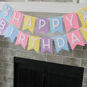 Puede incluir: Una pancarta de colores pastel con las palabras "Happy Birthday" en letras blancas. La pancarta est&aacute; decorada con recortes de conejos.