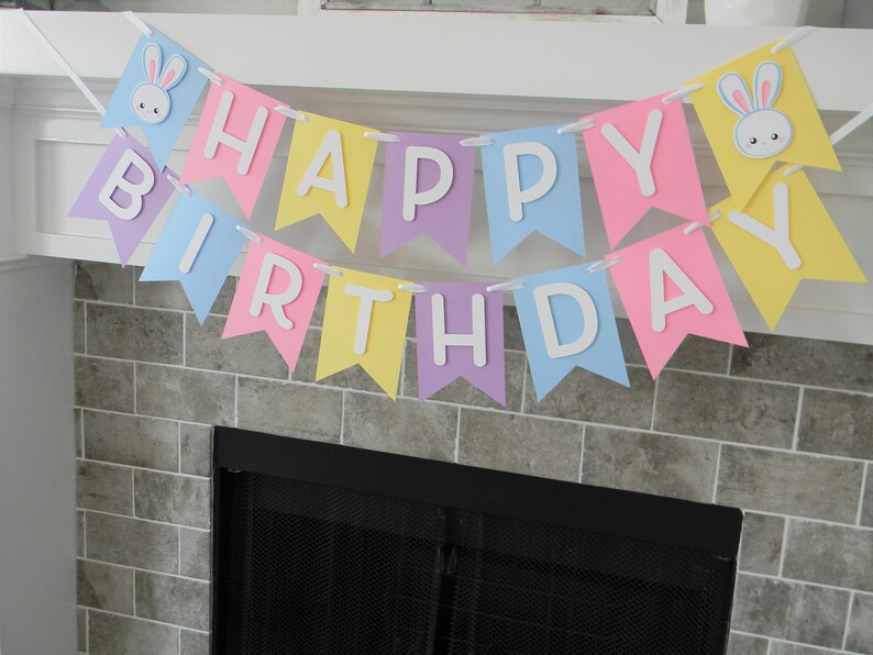 Puede incluir: Una pancarta de colores pastel con las palabras "Happy Birthday" en letras blancas. La pancarta est&aacute; decorada con recortes de conejos.