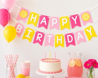 May include: A birthday party setup featuring a Happy Birthday banner in pink and yellow with lemon accents, a pink balloon, a yellow balloon, a white frosted cake with three candles, pink striped straws, pink cups, and a pitcher of pink lemonade.