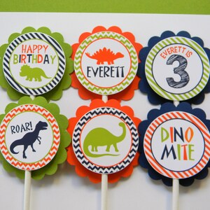 May include: Six green, orange and blue cupcake toppers with dinosaur designs. The toppers feature a variety of dinosaurs, including a triceratops, stegosaurus and tyrannosaurus rex. The toppers also include the text "Happy Birthday", "Everett", "Everett is 3", "Roar!" and "Dino Mite".