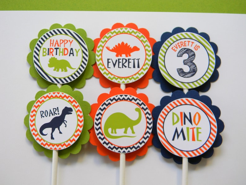 May include: Six green, orange and blue cupcake toppers with dinosaur designs. The toppers feature a variety of dinosaurs, including a triceratops, stegosaurus and tyrannosaurus rex. The toppers also include the text "Happy Birthday", "Everett", "Everett is 3", "Roar!" and "Dino Mite".