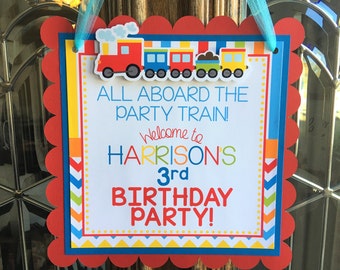 Op de afbeelding: Een kleurrijk trein-thema verjaardagsfeestbord met gegolfde randen. Het bord zegt "All aboard the party train! Welcome to Harrison's 3rd Birthday Party!"