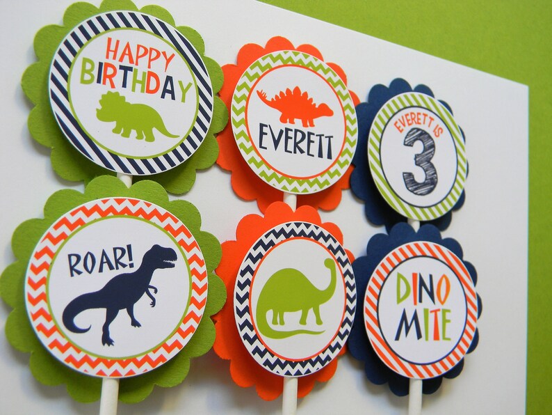 May include: Six green, orange and blue cupcake toppers with dinosaur illustrations and text. The toppers feature a green dinosaur, an orange dinosaur, a blue dinosaur, the words "Happy Birthday", "Roar!" and "Dino Mite", and the name "Everett" with the number "3".