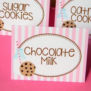 May include: Three pink and white striped signs with brown text and illustrations. The signs read "Sugar Cookies", "Oatmeal Cookies", and "Chocolate Milk". Each sign features a cartoon milk bottle with a straw and a chocolate chip cookie.