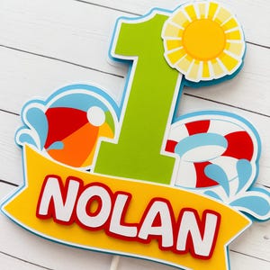 May include: A colorful cake topper with a beach theme. The number "1" is in green, with a yellow sun, a beach ball, and a life preserver. The name "NOLAN" is displayed on a yellow banner with red lettering.