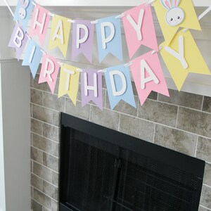 Puede incluir: Una pancarta de colores pastel con las palabras "Feliz cumplea&ntilde;os" y recortes de conejos. La pancarta est&aacute; colgada sobre una chimenea.