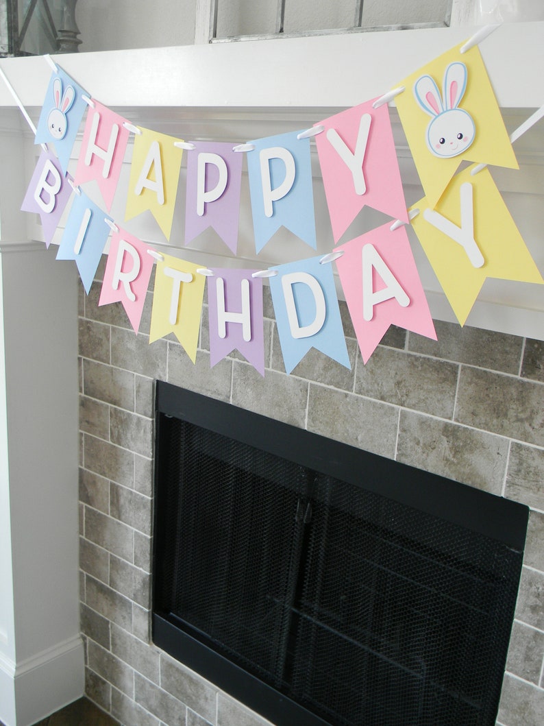 Puede incluir: Una pancarta de colores pastel con las palabras "Feliz cumplea&ntilde;os" y recortes de conejos. La pancarta est&aacute; colgada sobre una chimenea.