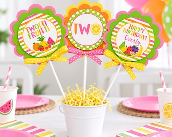 May include: Three decorative party centerpiece sticks in a white bucket filled with yellow paper shreds. The sticks feature scalloped edges, pink polka dot bows, and fruit-themed graphics with text reading Twotti Fruitti, Two, and Happy Birthday Everly.