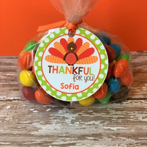 Op de afbeelding: Een doorzichtige plastic zak gevuld met kleurrijke snoepjes, vastgebonden met een groen-wit geblokt lint. Een rond label met een kalkoenillustratie en de tekst "THANKFUL for you! Sofia" is aan de zak bevestigd.