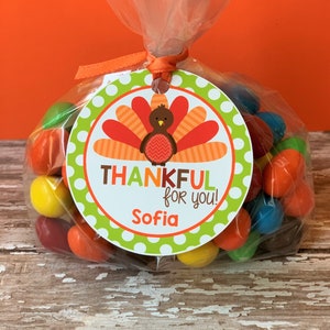 Op de afbeelding: Een ronde groene en witte cadeau-tag met een kalkoenillustratie en de tekst "Thankful for you!" en een naam "Sofia". De tag is bevestigd aan een doorzichtige plastic zak gevuld met kleurrijke snoepjes.
