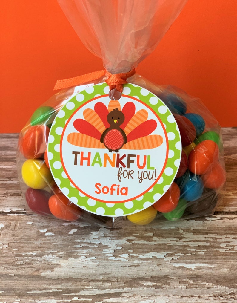 Op de afbeelding: Een ronde groene en witte cadeau-tag met een kalkoenillustratie en de tekst "Thankful for you!" en een naam "Sofia". De tag is bevestigd aan een doorzichtige plastic zak gevuld met kleurrijke snoepjes.