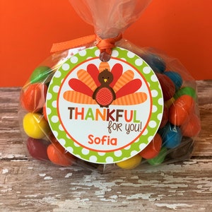 Op de afbeelding: Een groen en wit rond label met een kalkoenillustratie en de tekst "THANKFUL for you!" en de naam "Sofia". Het label is vastgemaakt aan een doorzichtige plastic zak gevuld met kleurrijke snoepjes.