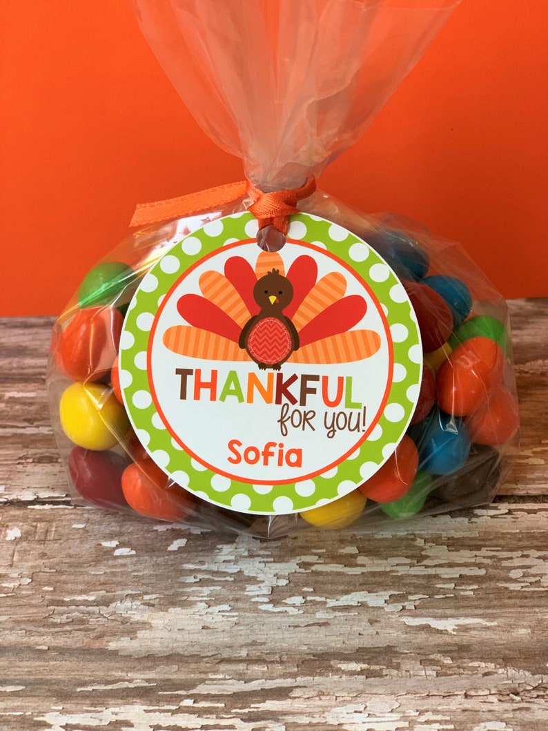 Op de afbeelding: Een groen en wit rond label met een kalkoenillustratie en de tekst "THANKFUL for you!" en de naam "Sofia". Het label is vastgemaakt aan een doorzichtige plastic zak gevuld met kleurrijke snoepjes.