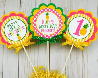 May include: Three yellow, pink, and green cupcake toppers with scalloped edges. The toppers feature a pineapple design and the text "Party Like a Pineapple", "Happy Birthday Penelope!" and "1".