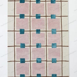 Puede incluir: Alfombra rectangular con fondo crema, patrón de cuadrícula con líneas marrones, cuadrados rosas y cuadrados turquesa en el centro. El diseño de la alfombra es moderno y geométrico, ideal para el hogar.