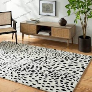 Puede incluir: Una alfombra rectangular, hecha a mano, con un fondo blanco y un patrón de manchas negras. La alfombra está colocada sobre un suelo de madera en una habitación con un mueble de madera, una silla y una planta en maceta. El diseño de la alfombra se asemeja al de un dálmata.