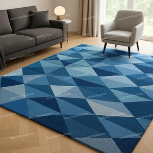 Puede incluir: Una alfombra rectangular grande con un patr&oacute;n geom&eacute;trico de tri&aacute;ngulos azules y celeste. La alfombra est&aacute; colocada sobre un suelo de madera, con un sof&aacute; gris, un sill&oacute;n y una peque&ntilde;a mesa auxiliar con una l&aacute;mpara en el fondo. Las dimensiones de la alfombra no son visibles.