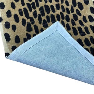 Puede incluir: Una alfombra con estampado de leopardo en beige y negro, con un reverso azul visible en la esquina. El patrón de la alfombra consiste en manchas negras sobre un fondo beige. La esquina está doblada.