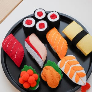Peut inclure: Un ensemble de sushis en feutrine sur une assiette noire. L'ensemble comprend des nigiris, des makis et d'autres variétés de sushis de différentes couleurs, dont le rouge, l'orange, le blanc et le noir. Les sushis sont disposés sur une assiette noire.