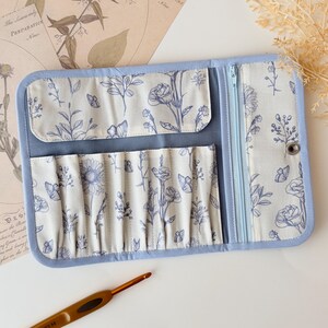 Peut inclure: Un étui de rangement en tissu bleu clair avec un intérieur crème orné d'un motif floral et de papillons bleus. L'étui comprend plusieurs fentes verticales pour le rangement, un compartiment zippé et une fermeture à bouton-pression en métal.