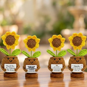 May include: Four crocheted sunflower ornaments. Each features a yellow flower with a brown centre, green leaves, and a brown pot with a smiling face. Each pot holds a sign with a different message, including "Thank You For Helping Me Grow."