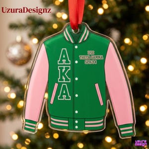 May include: A clear acrylic Christmas ornament shaped like a green and pink varsity jacket. The jacket features the letters AKA in white, along with the text "USC THETA GAMMA SPR 94". A red ribbon is attached for hanging.