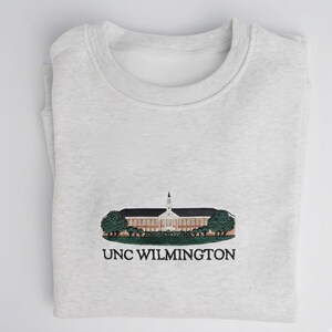 May include: A light gray sweatshirt with a crew neck. The front features an embroidered design of a building with a steeple, trees, and the text "UNC WILMINGTON" in black.