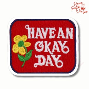 May include: Embroidered patch with a red background, blue border, and white text that reads "HAVE AN OKAY DAY." A yellow flower with a green stem and leaves is on the left. The patch is a rectangle with rounded corners.