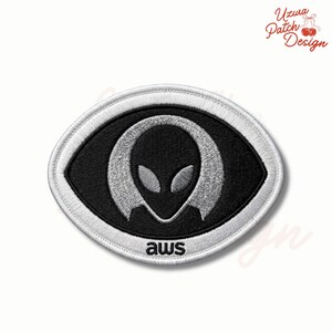 May include: Embroidered patch featuring a black alien head design within a black oval shape, outlined in white. The text "aws" is at the bottom. The patch has a white border and is likely for clothing or accessories.