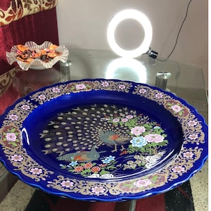 Puede incluir: Una fuente ovalada azul cobalto con un diseño de pavo real. La fuente presenta una ilustración detallada de dos pavos reales rodeados de flores rosas, azules y blancas. El borde está decorado con patrones florales dorados y rosas.