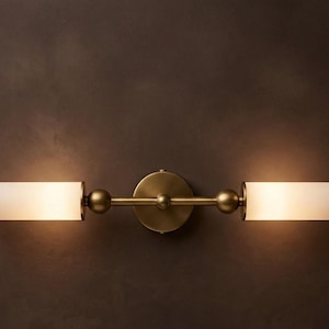 Può includere: Una lampada da parete con una barra orizzontale in metallo color ottone e una piastra di montaggio circolare centrale. Due paralumi cilindrici in vetro smerigliato si estendono dalle estremità, illuminati per emettere una luce calda.