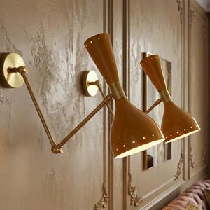 May include: Two mid-century modern wall sconces with a brass finish and tan shades. The sconces are mounted on a textured wall with decorative moulding. The shades have a conical shape with small perforations.