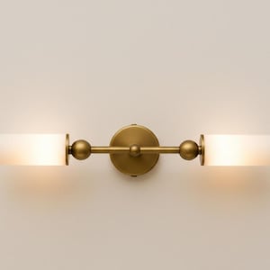 Könnte beinhalten: Messingfarbene Wandleuchte mit zwei zylindrischen, mattierten Glasschirmen. Die Leuchte hat eine zentrale kreisförmige Halterung, eine horizontale Stange und kugelförmige Akzente. Sie ist für die Wandmontage konzipiert und bietet Umgebungslicht. Ca. 40 cm breit.