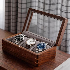 May include: A wooden watch box with a clear glass lid, holding three wristwatches. The watches have silver and blue faces, and leather and metal bands. The box is made of dark wood and has a gray velvet interior.