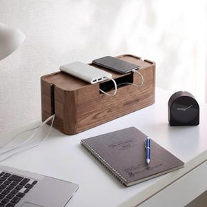 May include: A wooden cable management box with a power bank and smartphone charging inside. A black clock, notebook, and pen are on the white desk. The box has slots for cords to pass through.